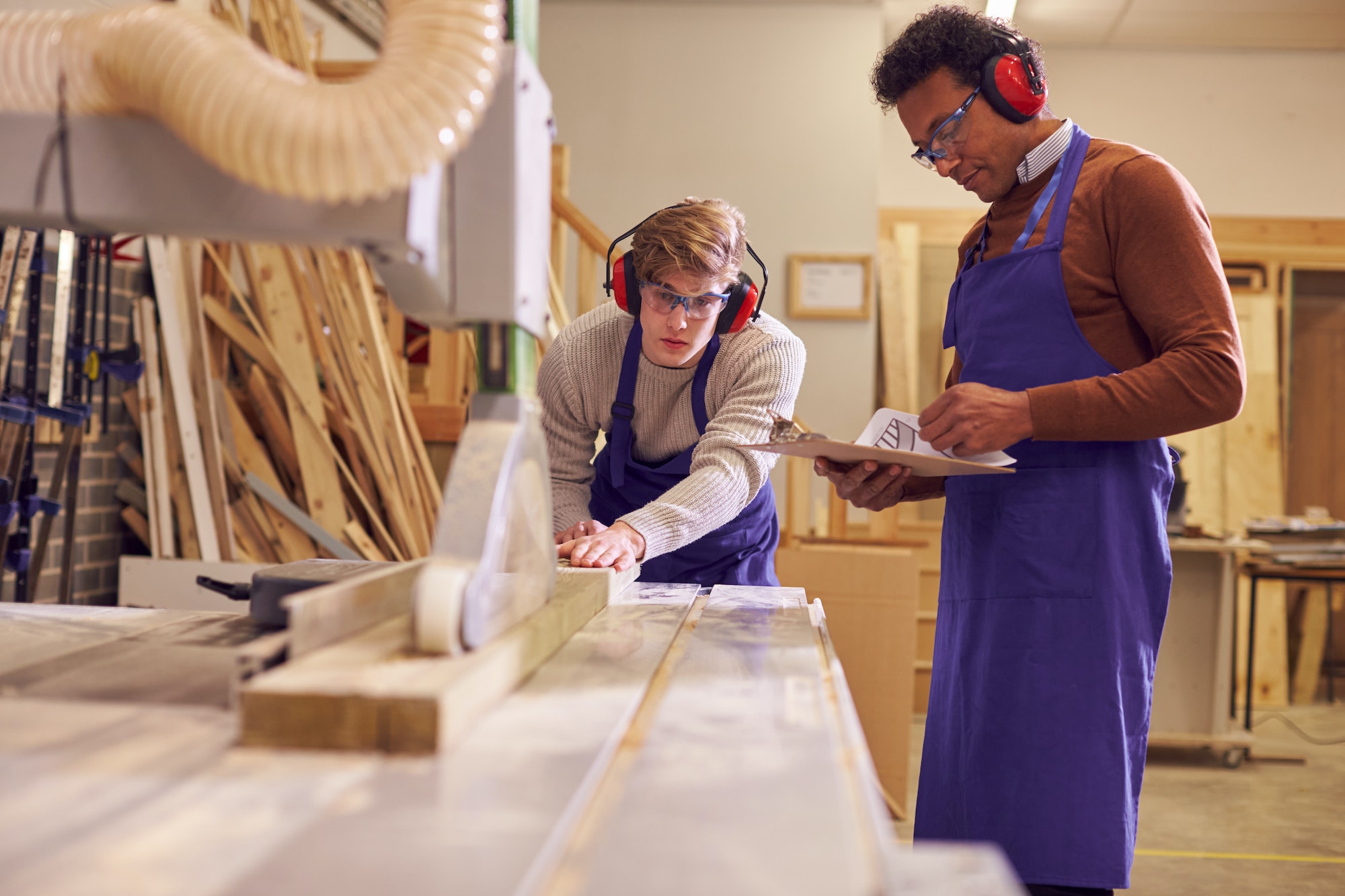 Tutor With Male Carpentry Student In Workshop Studying For Apprenticeship At College Using Bench Saw